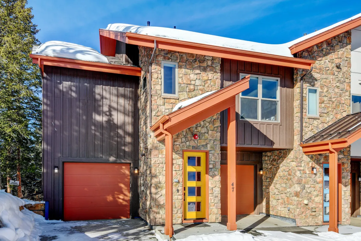 A welcoming exterior of a chalet with a warm stone facade, orange architectural accents, and a visible front entrance and garage. Snow blankets the ground, and soft lighting hints at cozy gatherings inside and easy access to outdoor adventures.