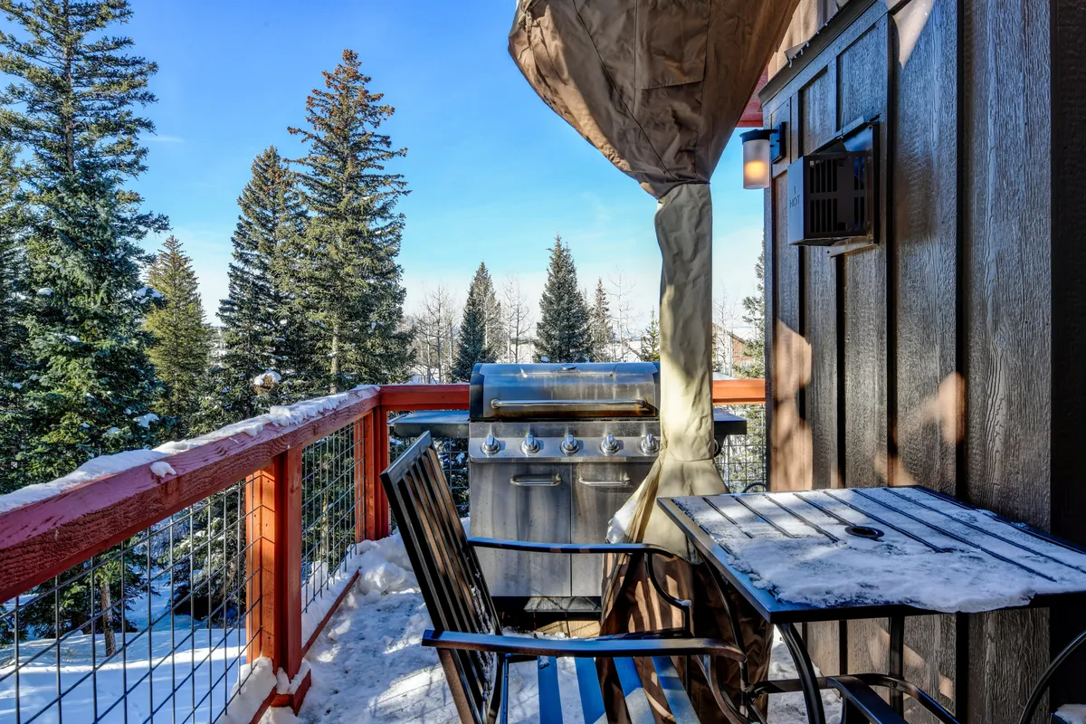 An inviting patio with a gas grill, table and chairs, and snow-covered railings, framed by evergreens. The setup suggests cozy outdoor meals, relaxation, and seamless access to nature right from the doorstep.