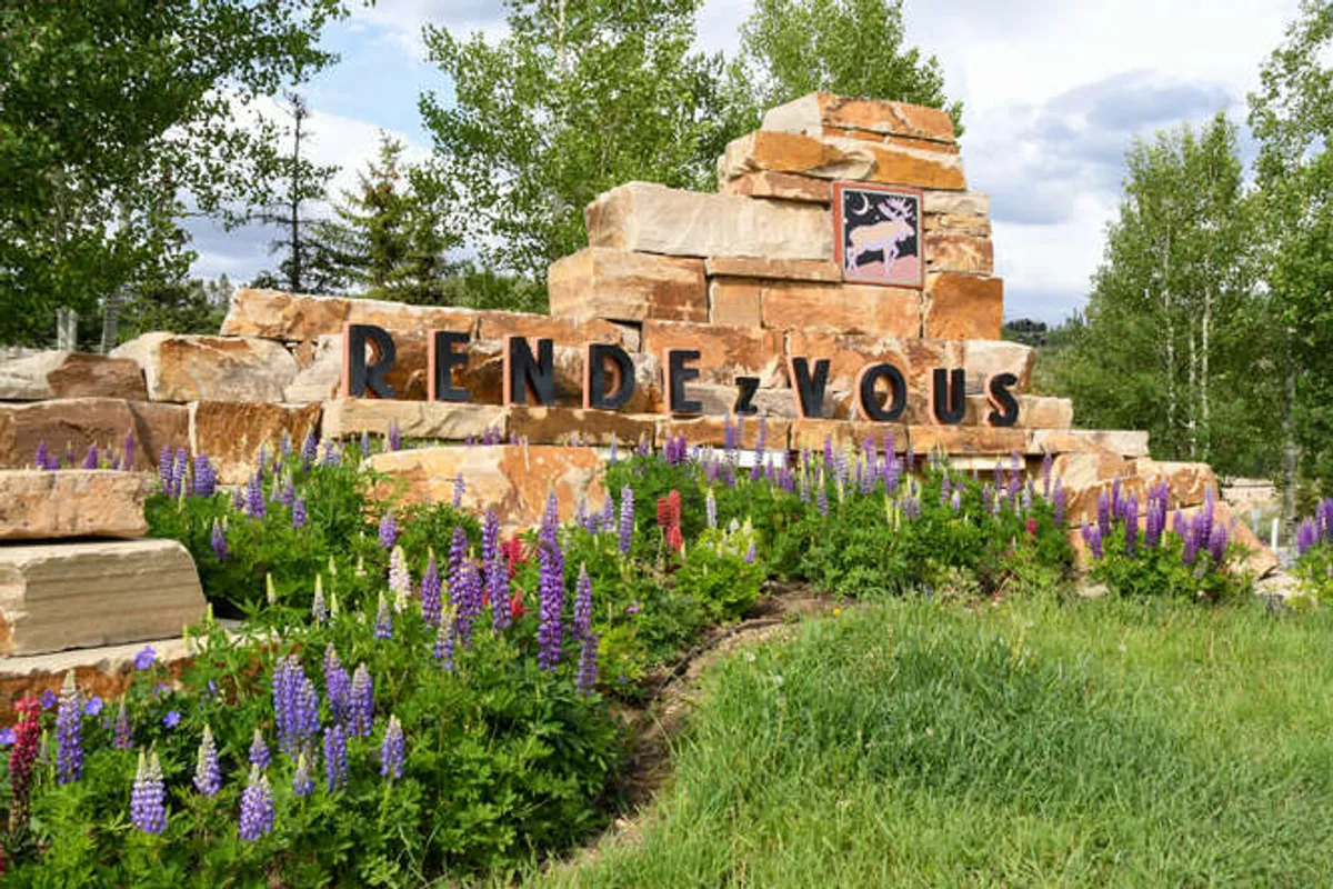 This image captures the stone monument reading 'RENDEZ-VOUS' surrounded by colorful flower beds and mature trees. The scene is bright, inviting, and highlights the neighborhood’s welcoming entrance and natural beauty.