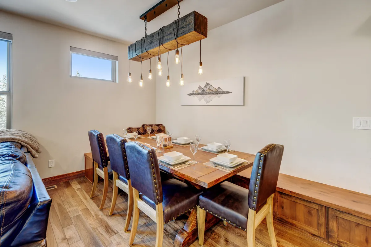 A warm and inviting dining area with a wooden table, hanging light fixture, and mountain artwork on the wall. The space is set for family dinners, celebrations, and morning coffee with panoramic views.