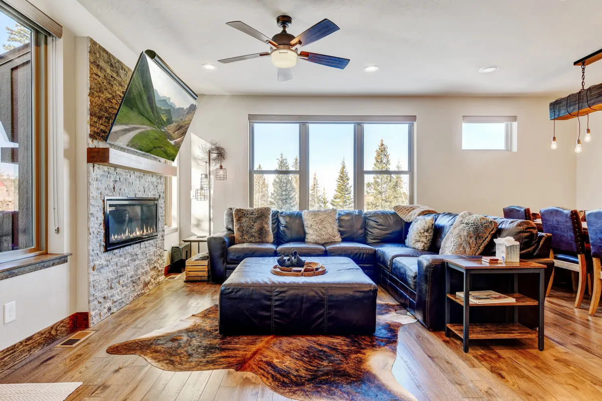A spacious, sunlit great room featuring a large sectional sofa, stone fireplace, mountain-view artwork, and a dining area with a wooden table. The open-concept layout invites both relaxation and entertainment, while oversized windows welcome in natural light and offer scenic views.