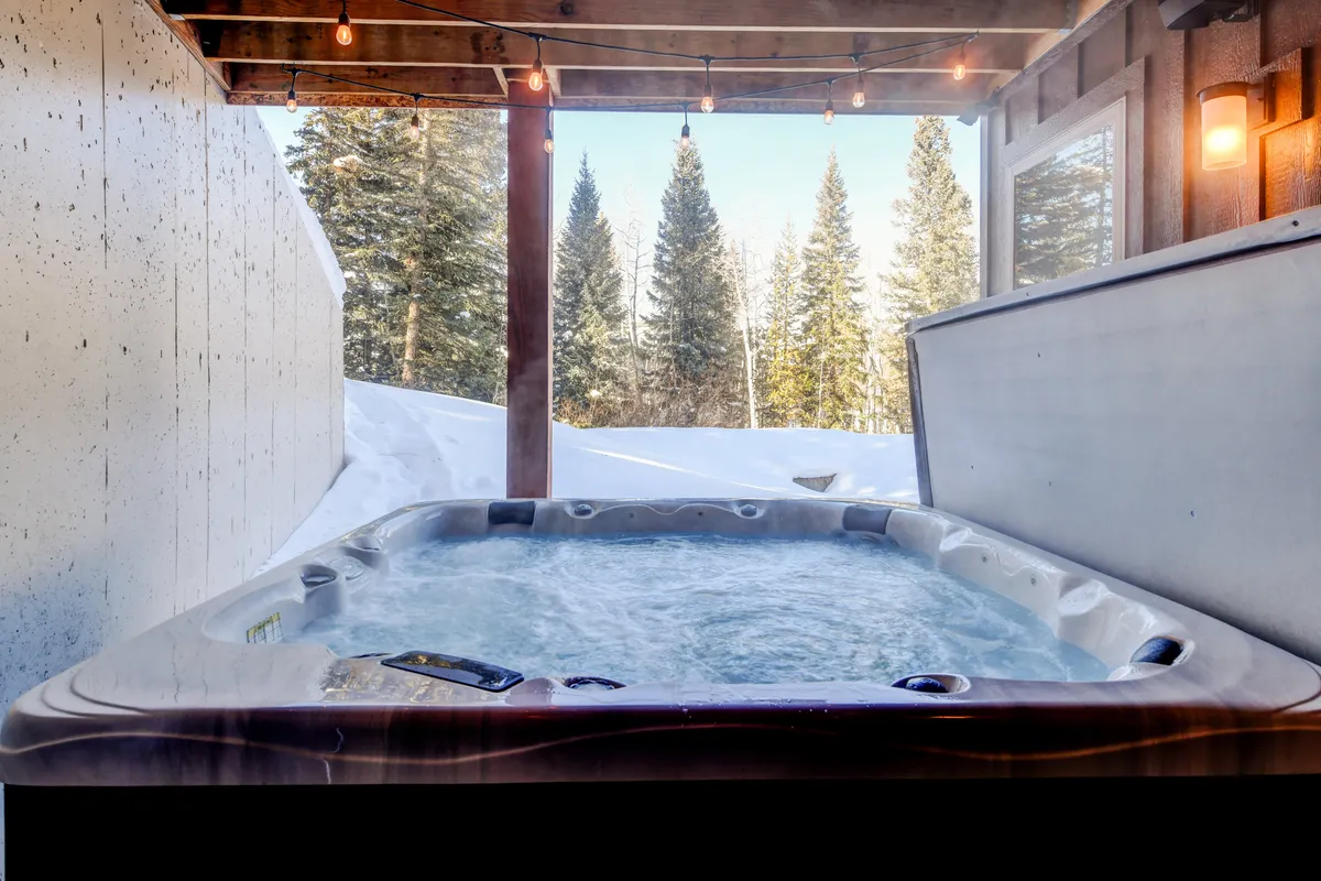 A steaming hot tub sits on a snow-blanketed deck, surrounded by pine trees and twinkling string lights. The backdrop of a tranquil winter landscape makes this outdoor amenity both a focal point for relaxation and a symbol of après-ski indulgence.