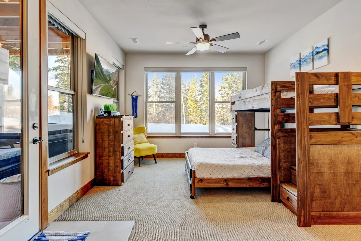 A cozy bunk room featuring a sturdy bunk bed, chest of drawers, and bright yellow accent chair. Large windows fill the space with natural light and offer views of nature, creating a cheerful, family-friendly atmosphere perfect for kids to play, read, or unwind after a day outdoors.