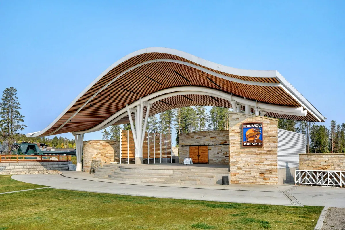 A dynamic exterior shot of a modern event center featuring a distinctive curved roof, stone facade, and clear event signage. The space is bustling with outdoor performances, cultural festivals, and community gatherings.