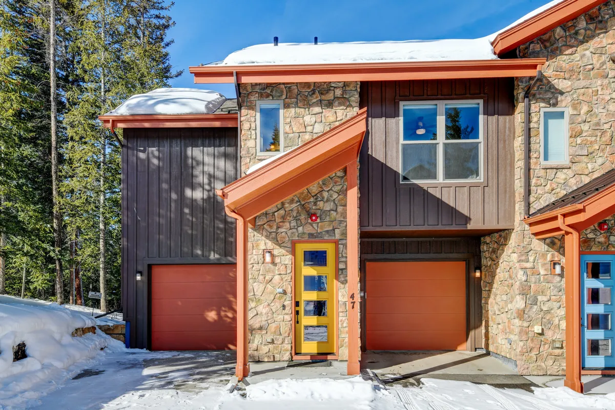 This image showcases the chalet's exterior with a welcoming stone facade, an inviting orange door, large windows, and garage doors. The snow-covered setting suggests year-round appeal—perfect for both winter sports and summer gatherings. The home's contemporary mountain design radiates warmth and comfort.