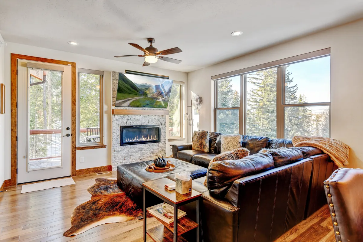 The image showcases a welcoming living room with a crackling stone fireplace as its centerpiece, surrounded by a plush leather sofa adorned with decorative pillows. Towering windows flood the space with natural light and reveal glimpses of Colorado's stunning mountain scenery. The décor is modern yet warm, inviting guests to relax, socialize, or unwind with a book while soaking in the views.